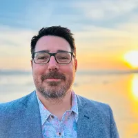 Carl Haggerty - Vice Chair - Carl looks at the camera smiling, wearing glasses and a suit, standing on the beach with the sun behind him